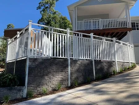 Stacked-stone concrete sleeper retaining wall below a white-railed deck beside a driveway