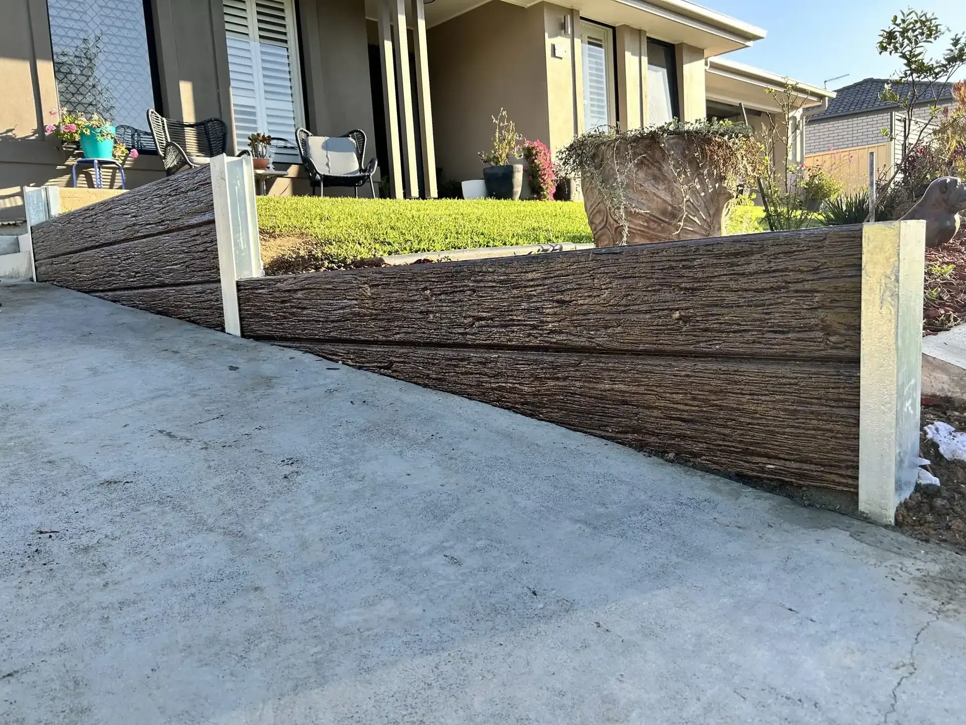 Woodgrain concrete sleeper retaining wall along a sloping driveway in front of a home