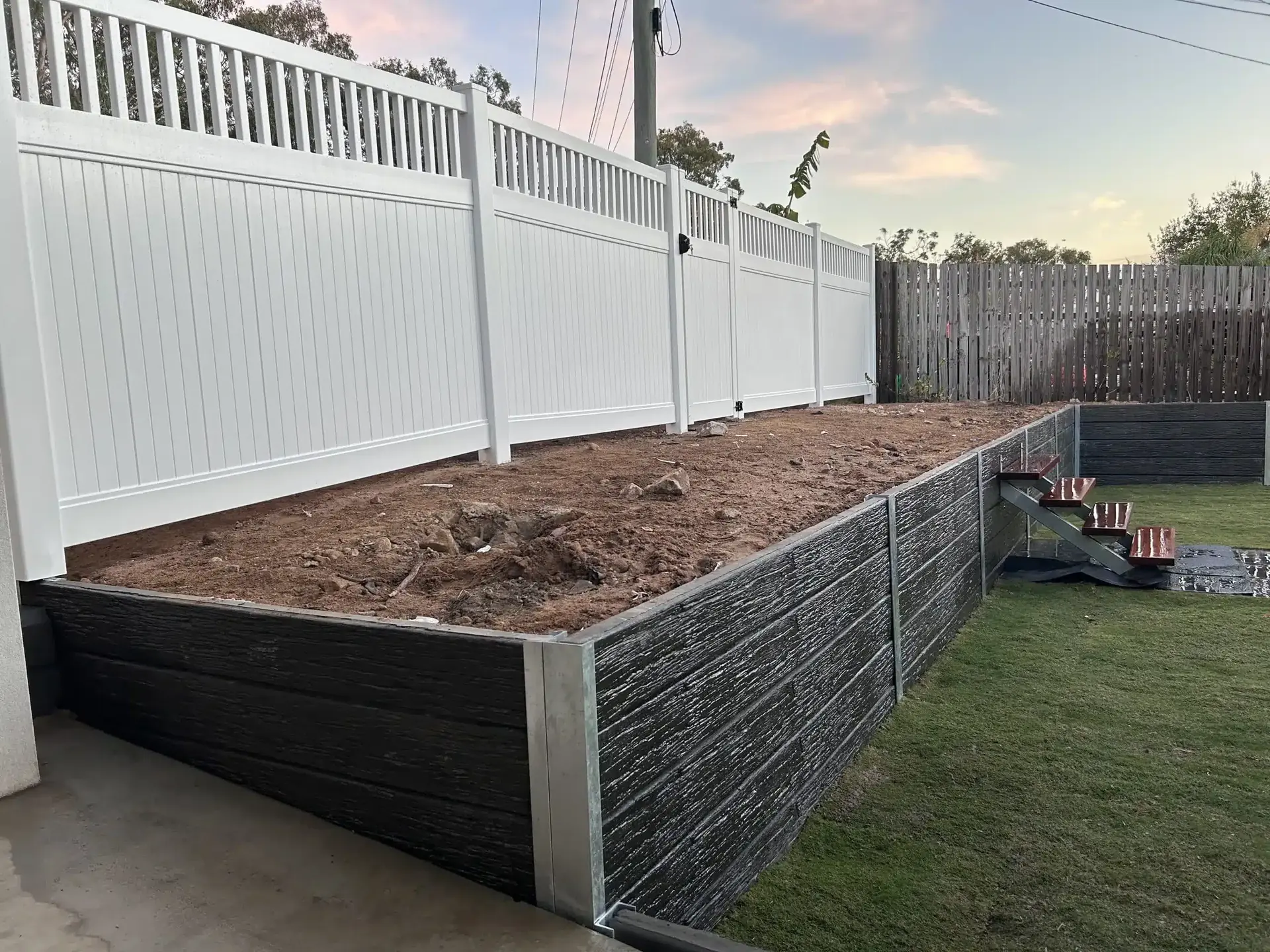 Charcoal concrete sleeper retaining wall around a raised garden area beside a white fence