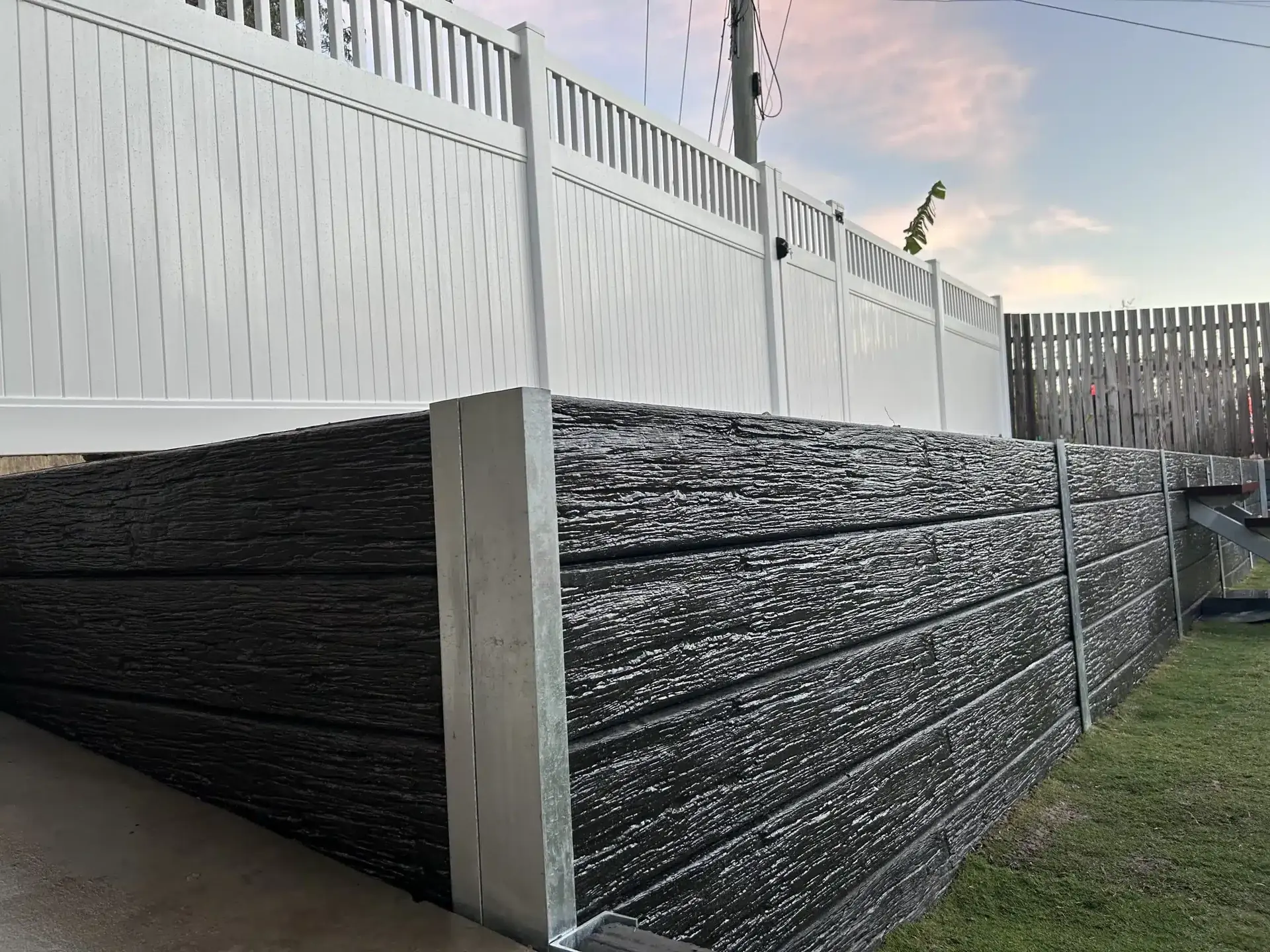 Charcoal woodgrain concrete sleeper retaining wall below a white vinyl fence