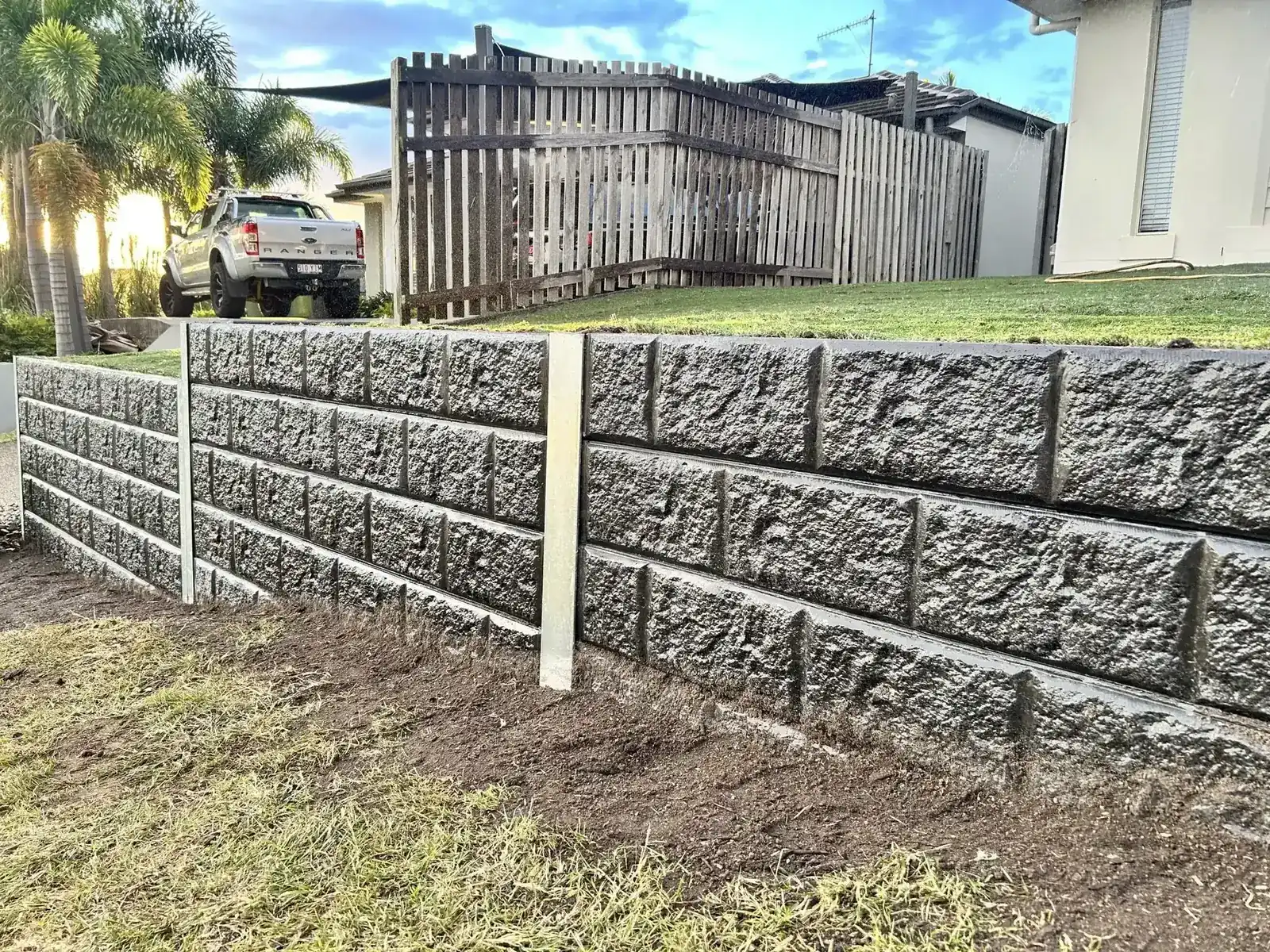 Grey rock-face concrete sleeper retaining wall with a steel post across a front yard