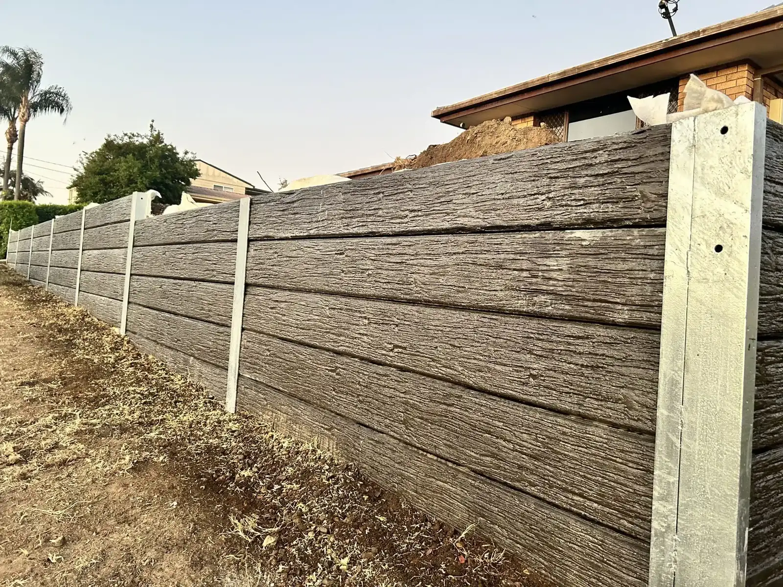 Woodgrain concrete sleeper retaining wall with galvanised posts along a sloping boundary