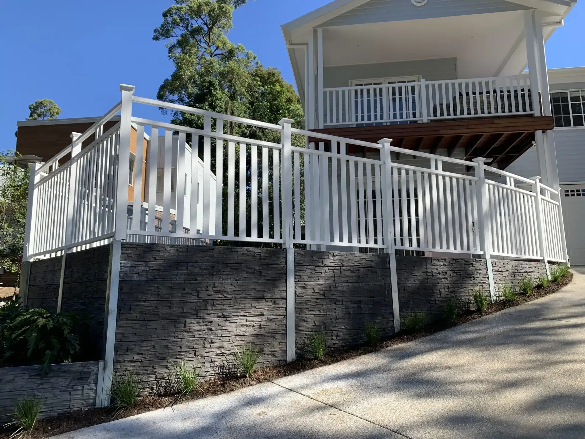 Stacked-stone concrete sleeper retaining wall below a white-railed deck beside a driveway