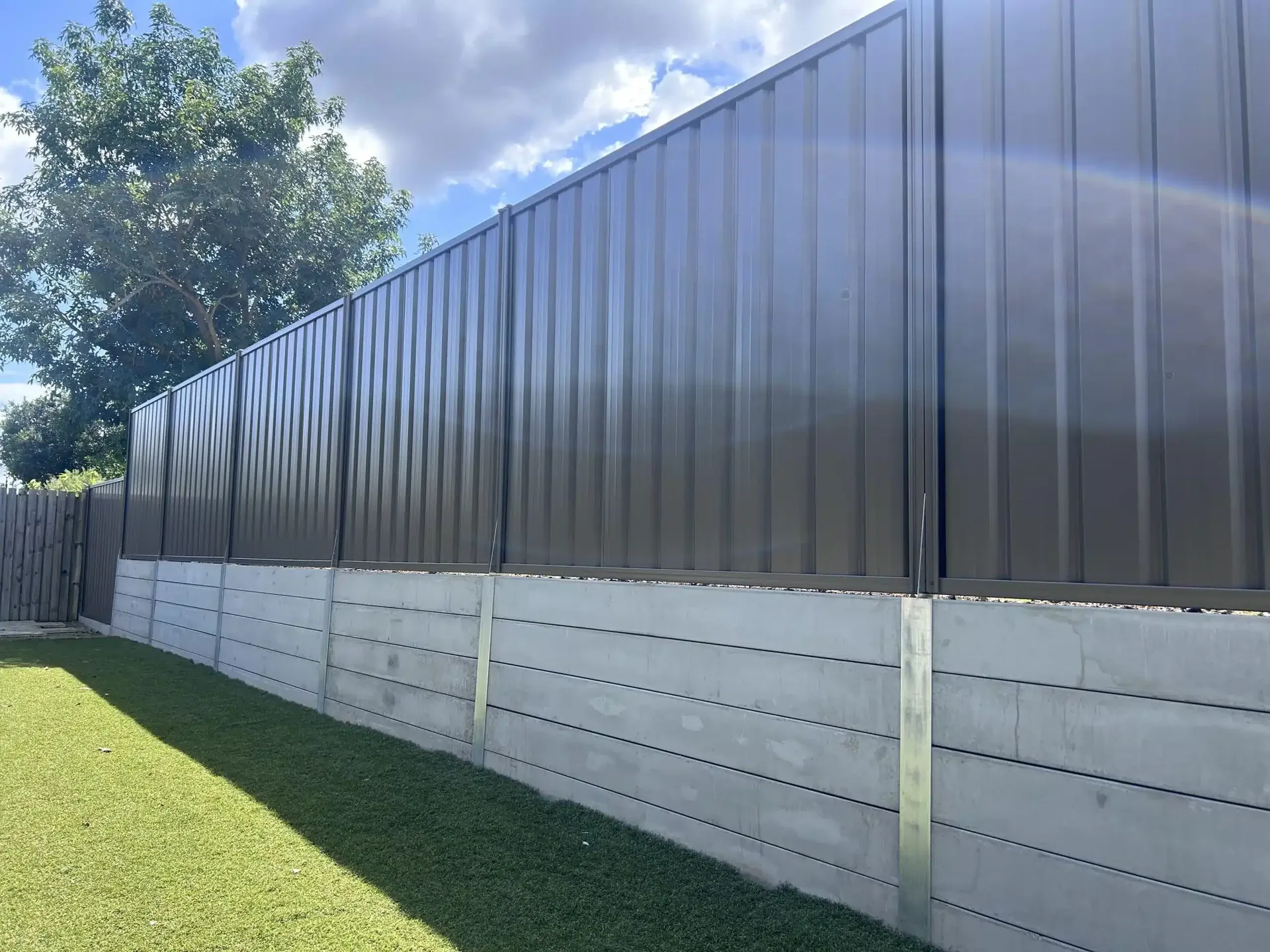 Smooth grey concrete sleeper retaining wall under a dark Colorbond fence beside a lawn