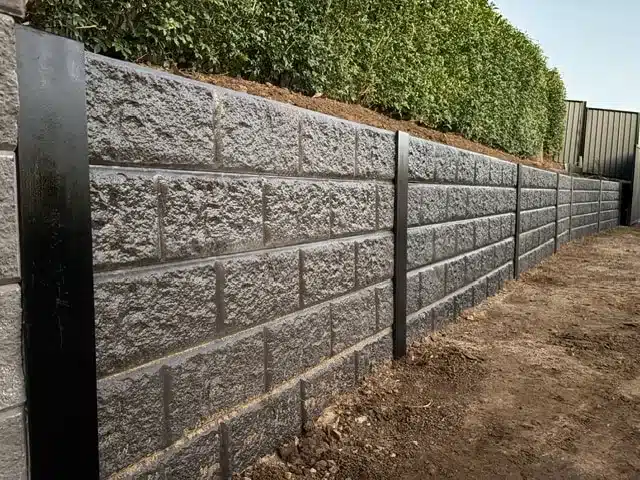 Charcoal block-pattern concrete sleeper retaining wall with black steel posts below a hedge