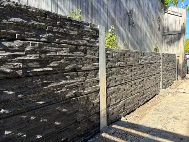 Dark stacked-stone concrete sleeper retaining wall with steel posts along a side boundary